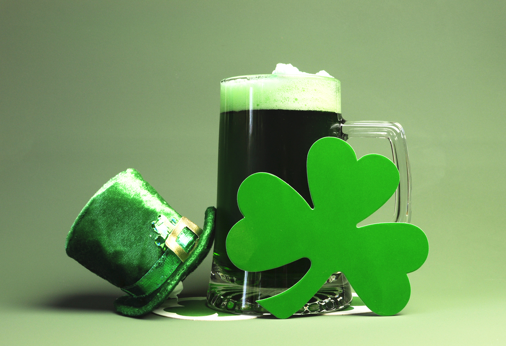 St. Patrick's Day - 3 leaf shamrock in front of glass mug of green beer with foam, sitting on two white coasters with green shamrocks, with a small green top hat on the left leaning against the glass 