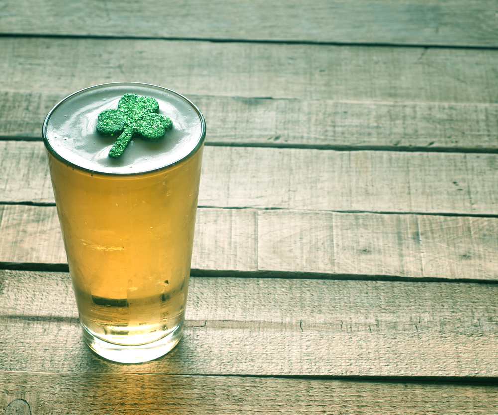 frosted glass of light amber beer with white foam and a glittery shamrock sitting on the foam against a background of a worn wooden deck with uneven planks