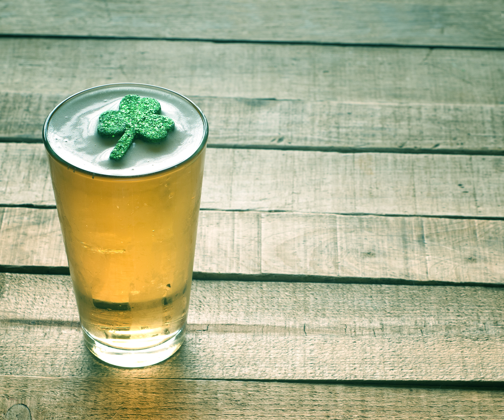 frosted glass of light amber beer with white foam and a glittery shamrock sitting on the foam against a background of a worn wooden deck with uneven planks frosted glass of light amber beer with white foam and a glittery shamrock sitting on the foam against a background of a worn wooden deck with uneven planks