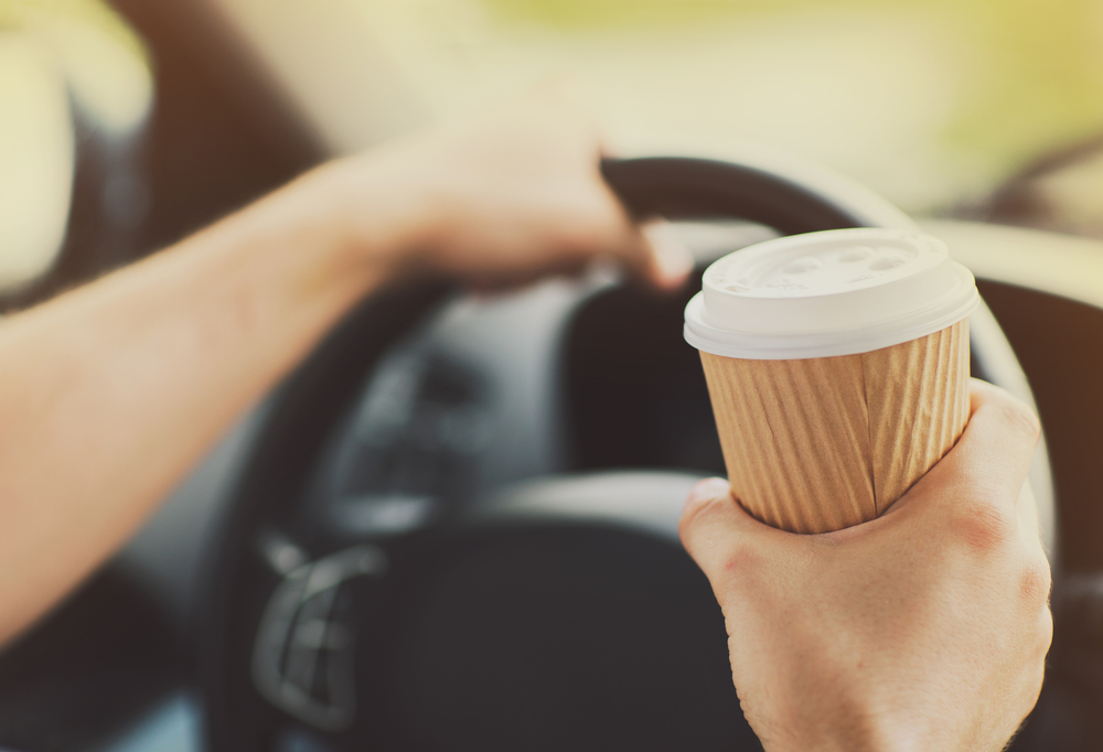 Driver holding paper cup of coffee in one hand and steering with the other hand