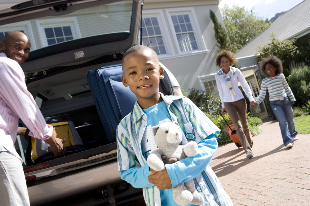 Boy smiling and holding stuffed animal with smiling father loading trunk of car and smiling mother roling suitcase, holding smiling sister's hand, walking together towards the car, all in front of house