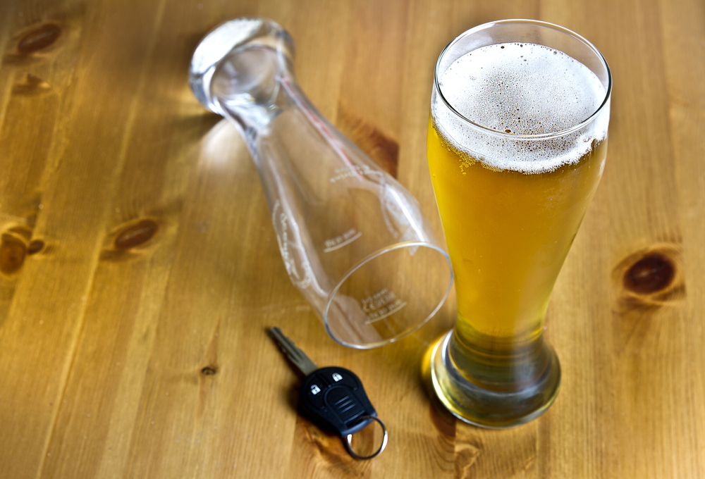 Fresh glass of light amber beer next to a car key and an empty, knocked over glass on a light wood table