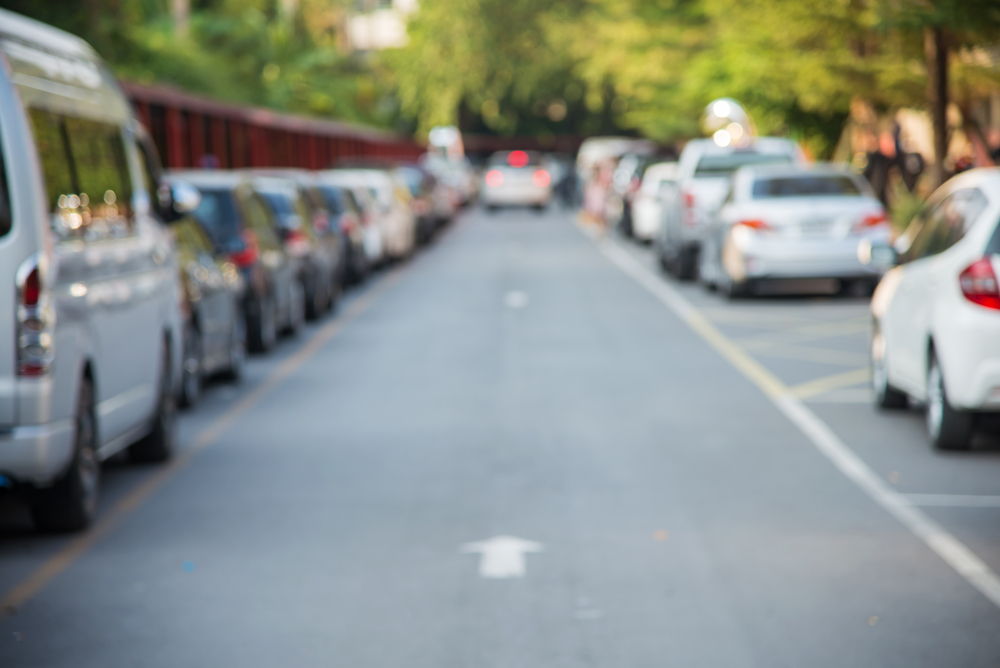 Blurred picture of car driving down street full of cars parallel parked on both sides of the street with beautiful trees lining the street on the right and a fence on the left