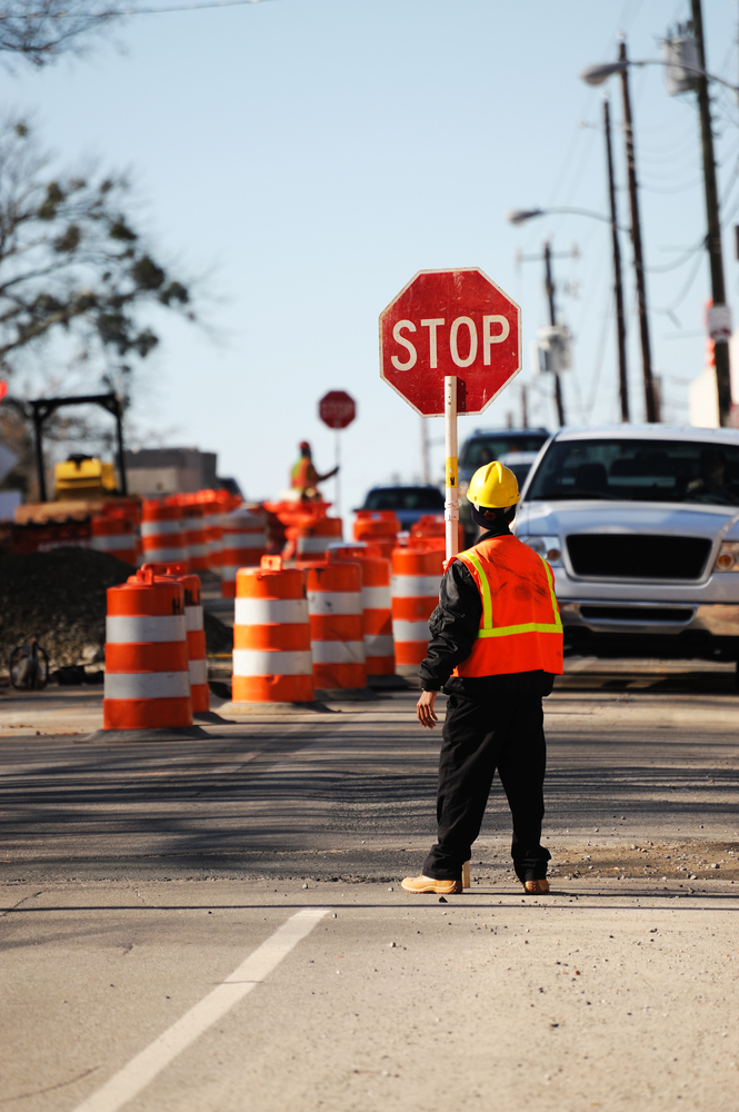Road construction worker in yellow hardhat and orange vest stopping traffic next to road work with bulldozers and orange barricades
